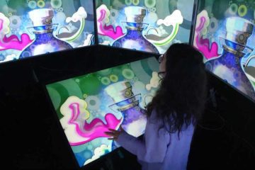 The image shows a person is interacting with a digital display inside an immersive exhibit at MADS Art Gallery. The screen depicts a vibrant and colourful digital art, depicting a stylized perfume bottle with a figure inside it. The bottle is primarily in hues of purple and blue, with an ethereal quality emphasized by surrounding swirling clouds and vapour-like elements in white and pink.. Multiple layers of the screen surrounding the person show repeating images of the same stylized perfume bottle shown on the main screen. The person appears to be touching or gesturing toward the display, engaged in an interactive component of the exhibit.