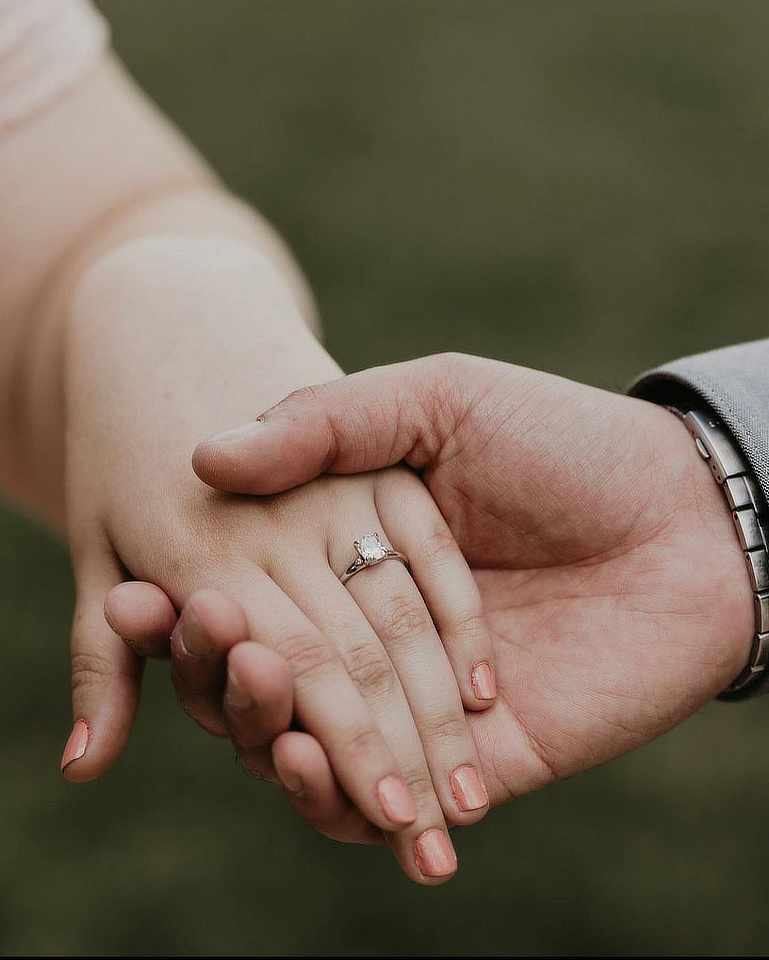 The image shows a close-up of two hands gently holding each other. One hand, with neatly manicured nails painted a soft peach color, wears an engagement ring featuring a prominent solitaire diamond. The other hand, appearing larger, is clasping it softly, with part of a wristwatch visible on the wrist. The background is out of focus, showing a muted green, likely grass or foliage, creating a serene and intimate feel.