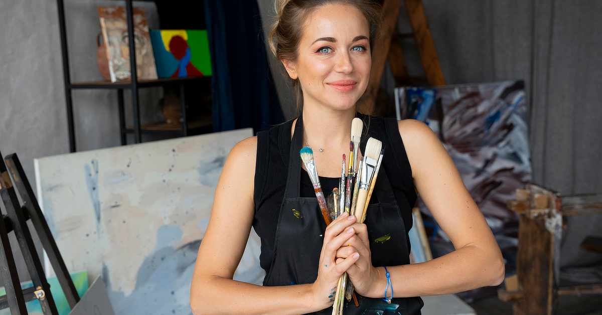 A woman wearing a black apron stands in an art studio, holding a bundle of paintbrushes in both hands. She has light brown hair tied up and is smiling slightly. Behind her are several abstract and colorful paintings, an easel, a wooden ladder, and shelving with more artwork. The setting appears to be a creative workspace for painting.