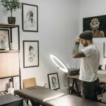 Interior of a tattoo studio with white walls covered in framed black-and-white artwork. A tattoo artist wearing a beanie and white T-shirt stands near a lit ring light, adjusting his hat. In the foreground is a black tattoo chair. Wooden shelves display plants, jars, and art prints, while a desk along the wall holds equipment and decorative items.