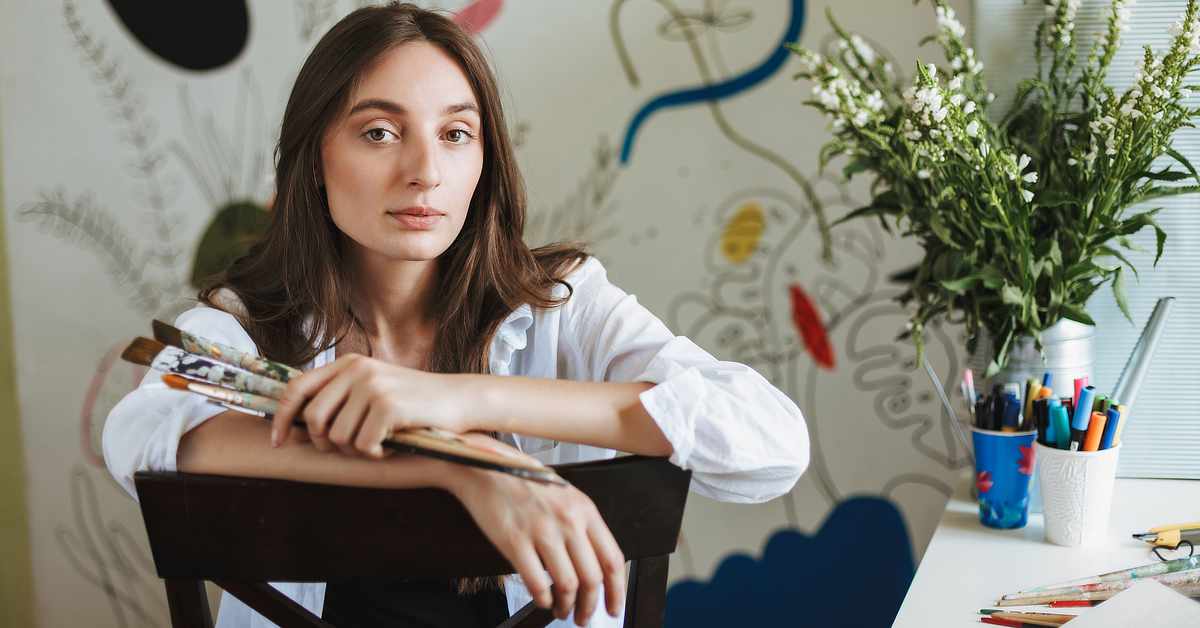 A person with long brown hair wearing a white button-up shirt leans on the back of a dark wooden chair while holding several paintbrushes. They face the camera with a neutral expression. In the background, a wall features abstract painted shapes and lines. To the right, a white desk holds cups filled with colorful markers and pens, a watering can, scattered art supplies, and a vase of white flowers near a window.