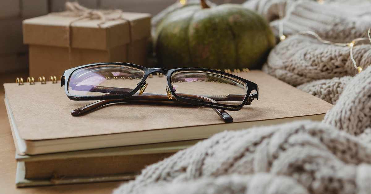 Black rectangular eyeglasses rest on a spiral notebook atop stacked books, with a gift box, green pumpkin, and knitted blanket softly blurred in the background.