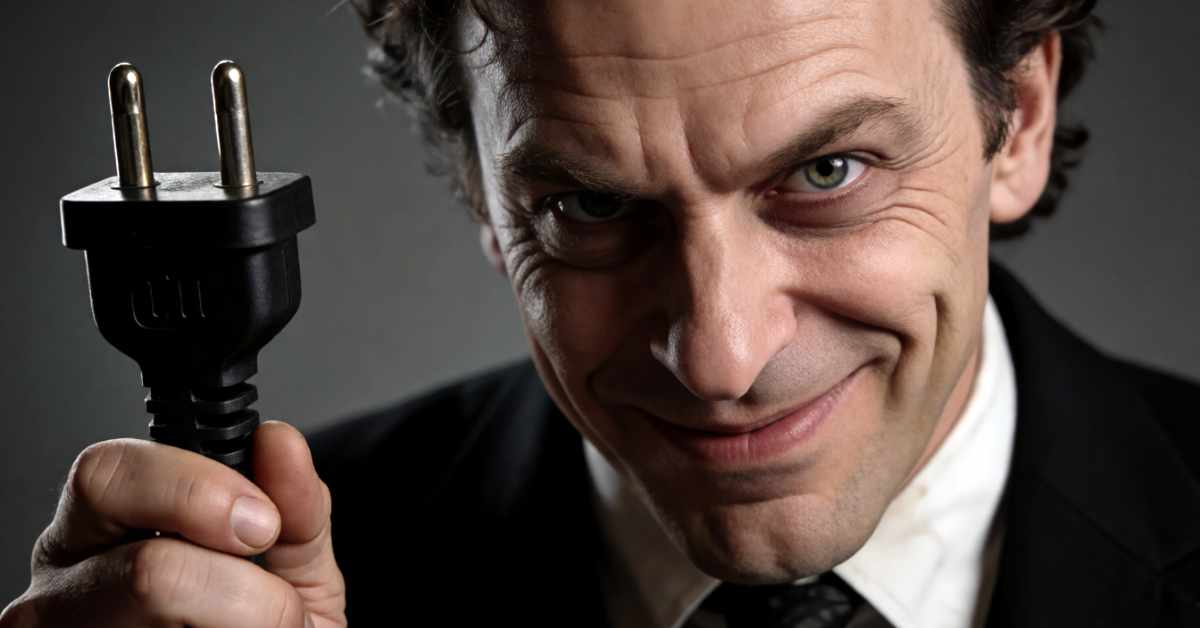 Close-up of a smirking man in a black suit holding a black two-prong electrical plug toward the camera.